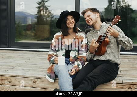 Hipster man playing on ukulele for his stylish woman, relaxing on wooden porch of modern cabin with big windows in mountains. Happy young family trave Stock Photo