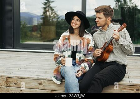 Hipster man playing on ukulele for his beautiful stylish woman, relaxing on wooden porch on background of modern cabin with big windows in mountains. Stock Photo