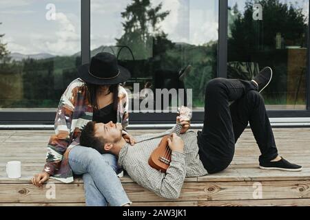 Hipster man playing on ukulele for his beautiful stylish woman, relaxing on wooden porch on background of modern cabin with big windows in mountains. Stock Photo