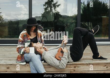 Hipster man playing on ukulele for his beautiful stylish woman, relaxing on wooden porch on background of modern cabin with big windows in mountains. Stock Photo