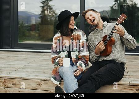 Happy young family travelers smiling and having fun. Hipster man playing on ukulele for his stylish woman, relaxing on wooden porch  of modern cabin w Stock Photo