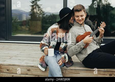 Happy hipster couple relaxing on wooden porch of modern cabin with big windows in mountains. Hipster man playing on ukulele for his stylish woman, enj Stock Photo