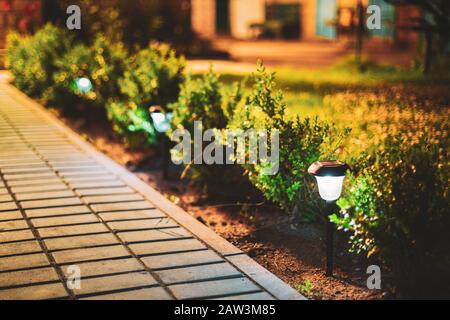 Night View Of Flowerbed Illuminated By Energy-Saving Solar Powered Lanterns Along The Path Causeway On Courtyard Going To House Stock Photo