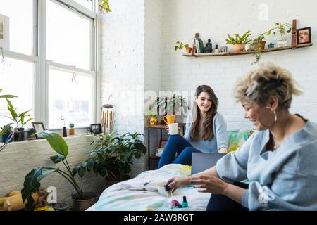 Woman with coffee mug using laptop while friend applying nail polish on bed at home Stock Photo