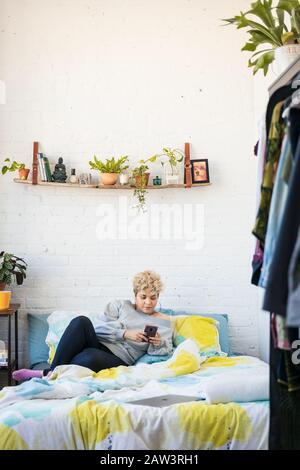 Woman using smart phone while leaning on bed against wall in bedroom at home Stock Photo
