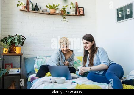 Woman showing laptop to roommate having coffee while sitting on bed at home Stock Photo