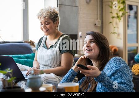 Happy young woman watching TV while friend using laptop at home Stock Photo