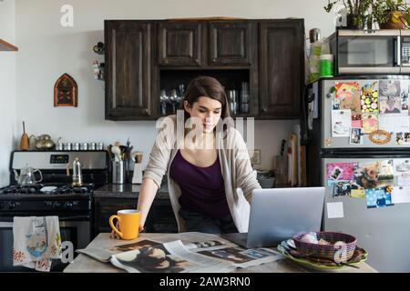 Young woman using laptop while having coffee at table in kitchen Stock Photo