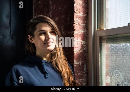 Thoughtful smiling young woman looking through window at home Stock Photo