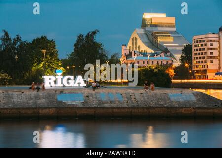 Riga, Latvia - June 30, 2016: Sitting People On Edge Of Urban Concrete Embankment Of Daugava River Around The Glowing City Name Sign In Summer Evening Stock Photo