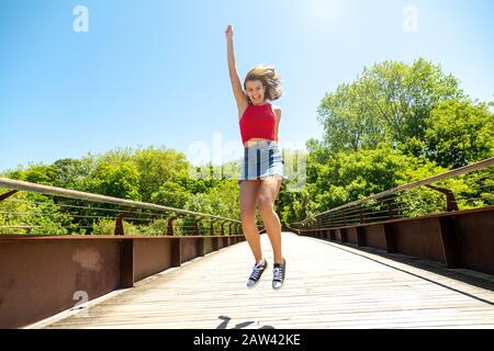 Happy young beautiful woman in red top and mini skirt jumping on a bridge in a sunny day Stock Photo