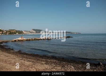 A lone female is sat relaxing on the rocks reading a book on a warm sunny day at a empty beach on Zakynthos island Greece. Stock Photo