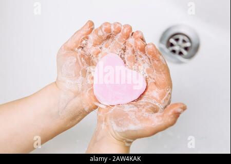 Importance of personal hygiene care. Flat lay view of child washing dirty hands with pink heart shape soap bar, lot of foam. Copy space. Stock Photo