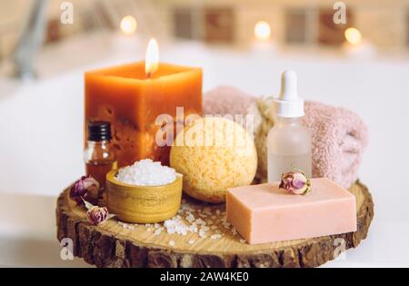 Home spa products on wooden disc tray: bar of soap, bath bomb, aroma bath salt, essential and massage oils, candle burning, towel inside bathroom. Stock Photo