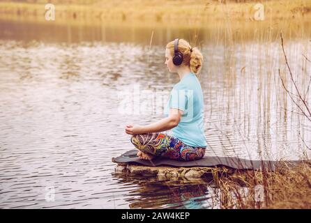 Blond hair woman sit outdoors by water and listen audio guided meditation from smartphone, wearing blue T-shirt sports clothing. Warm sunny day. Stock Photo