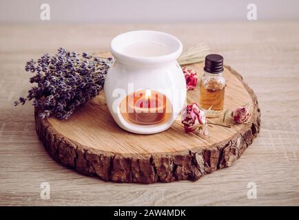 Vintage style picture of white ceramic candle aroma oil lamp with essential oil bottle and dry flower petals on natural pine wood disc, dry background Stock Photo