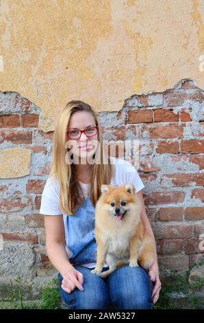 Young, cute next door girl wearing red glasses, jeans and t-shirt, posing with her pet in front of grunge brick wall Stock Photo