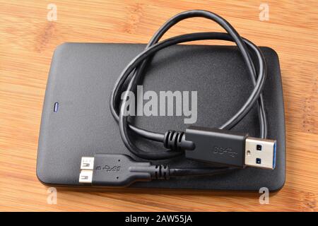 Black and flat external USB 3.0 hard disc with coiled USB 3.0 cable on wooden background, view from above Stock Photo