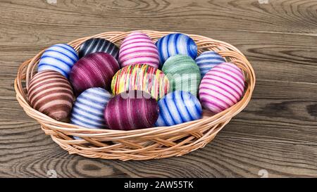 Colored striped ornate Easter eggs in wicker basket on wood. Empty eggshells hand-decorated by glued colorful cotton sewing yarn. Egg shell decoration. Stock Photo