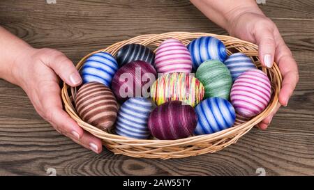 Female hands holding Easter eggs in wicker basket on wooden table detail. Ornate empty eggshells wrapped in colorful cotton thread. Holiday decoration. Stock Photo
