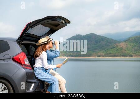 Young Asian couple with man tourist looking through binoculars while his smiling girlfriend sitting near with map on car trunk in rural field view wit Stock Photo