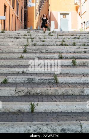 From below of young woman in casual black dress walking down stairs in city street in sunny summer day Stock Photo