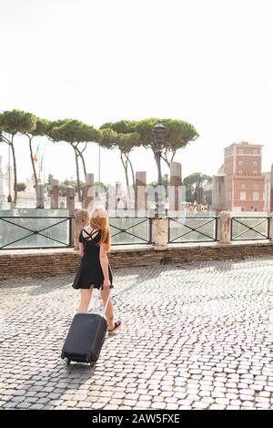 Back view of modern redhead female traveler in casual black dress pulling suitcase while walking on paved square in city during summer holidays Stock Photo