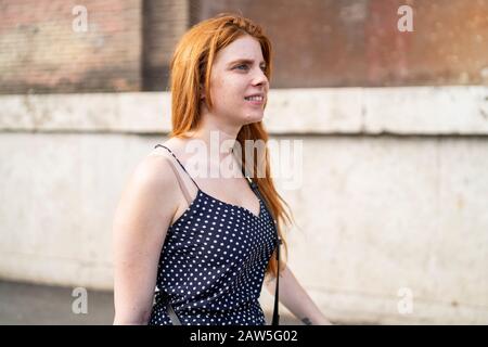 Positive young female with red hair smiling and walking outside shabby building on city street Stock Photo