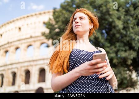 Low angle of pensive young lady with red hair browsing smartphone and looking away while visiting Colosseum on sunny day Stock Photo