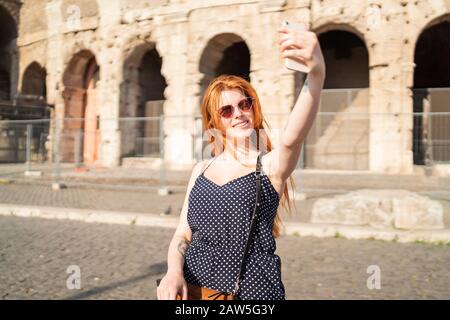 Delighted young woman in stylish outfit smiling and taking selfie while standing on pavement outside ancient Colosseum building Stock Photo