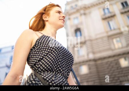 From below happy young woman with ginger hair smiling and walking on city street on sunny summer day Stock Photo
