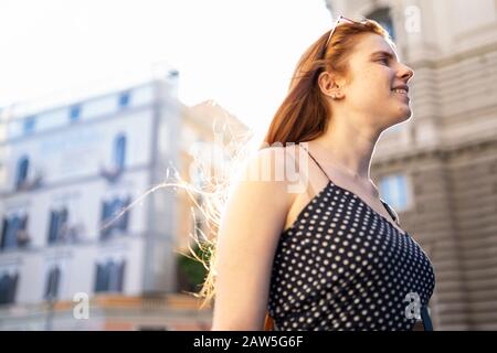 Low angle of optimistic young lady with long ginger hair smiling and enjoying sunny day while walking on city street in summer Stock Photo