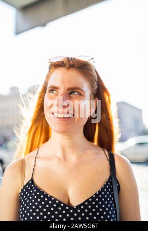 Positive young woman with red hair smiling and looking away while standing on city street on sunny day Stock Photo