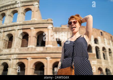 Low angle of trendy young female showing tongue and doing ginger hair while visiting Colosseum on sunny day in city Stock Photo