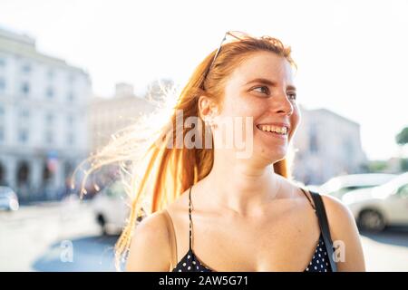 Gleeful young female with red hair smiling and looking away while standing on city street on sunny summer day Stock Photo