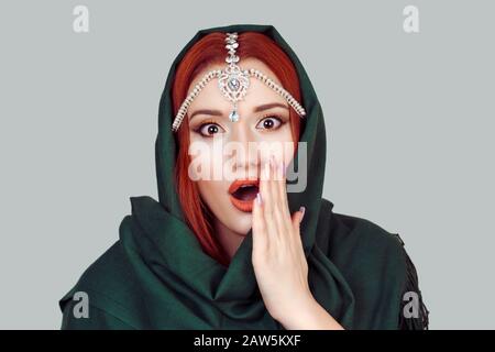 Beautiful shocked surprised muslim woman girl with big eyes with hand on open month looking at camera green hijab scarf and tikka jewelry on head isol Stock Photo