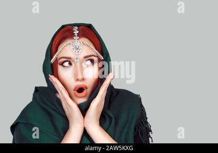 Shocked beautiful woman with big eyes, open month, with hand on cheeks looking to the side, side wards green hijab scarf and tikka jewelry on head iso Stock Photo