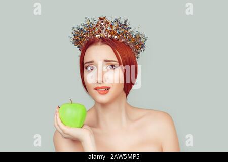 Beauty queen craving for vitamins sugars. Pretty woman with crystal crown on head holding green apple in hand biting lips isolated on light green gray Stock Photo