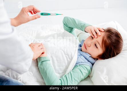 Sick child with flu fever laying in bed and mother holding thermometer Stock Photo