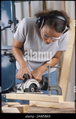 African-American young woman wears safety goggles and ear protection while making cut with cordless circular saw in her garage workshop in Austin, Texas. Stock Photo