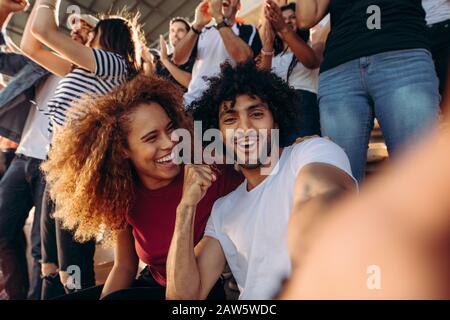 Couple of spectators taking selfie while cheering their sports team from a stadium. Cheerful man and woman fans taking selfie while watching match in Stock Photo