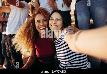 Crowd on a stadium watching a sports event taking selfie. Two women taking a selfie in stadium. Stock Photo