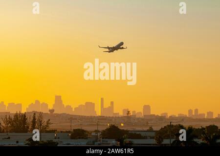 A plane is silhouetted against a colorful Yellow and Orange sunrise as it takes off from the Miami airport with the city in the background. Stock Photo