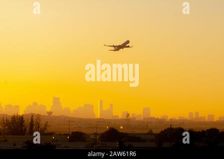 A plane is silhouetted against a colorful Yellow and Orange sunrise as it takes off from the Miami airport with the city in the background. Stock Photo