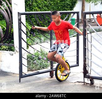 A young Asian boy, aged 12-13 years, practices riding a monocycle, initially holding on to a metal gate for support to steady himself, in Bangkok, Thailand, South East Asia. Stock Photo