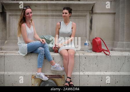 Two well dressed young Parisian women sit talking on a wide stone ledge of a grand architectural place at, Parvis Notre-Dame - Pl. Jean-Paul II. Stock Photo