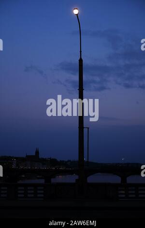 A lamp post on a Prague bridge shines against the night sky, everything else looking small scale and toy like. Stock Photo
