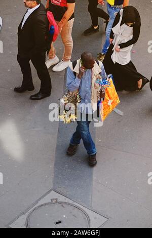 A street vendor souvenir seller at Bir-Hakeim metro stands with arms full of miniature Eiffel Tower souvenirs as people pass behind, viewed from above Stock Photo