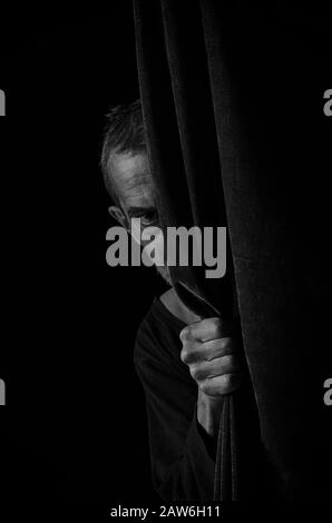A frightened man peeks out from behind a curtain in black and white. Stock Photo