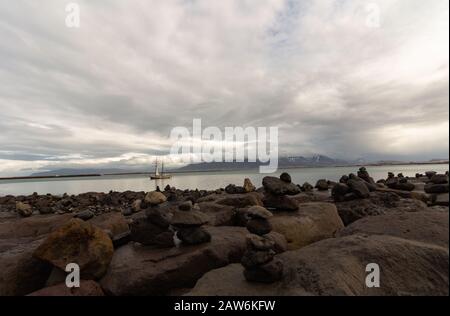 Along the pier next to the Art Museum in Reykjavik Iceland. Sailboats pass by while people balance or stack rocks, by the coastline. Stock Photo